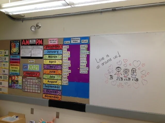Classroom wall with colorful educational posters, a calendar, a schedule of the months, and a handwritten note with drawings of children and hearts that says 'Love is all around us!'