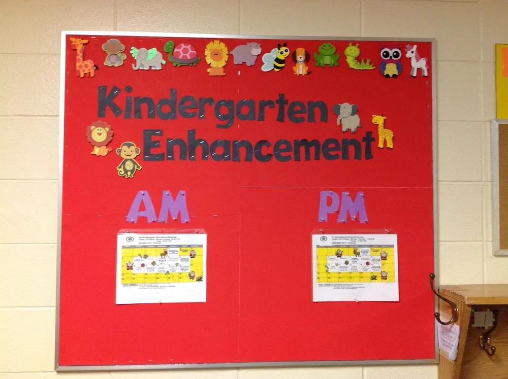 Red classroom bulletin board titled "Kindergarten Enhancement" decorated with animal cutouts at the top, and sections labeled "AM" and "PM" each with schedule sheets.