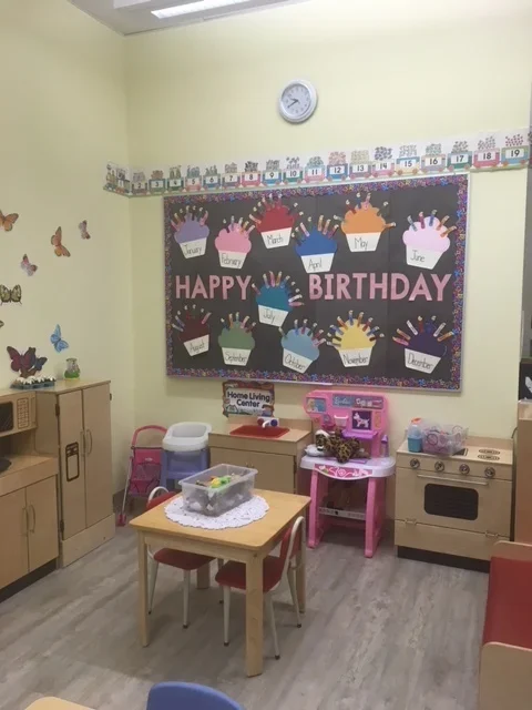 Children's birthday celebration room with decorated blackboard featuring colorful cupcake cutouts for each month, a clock on the wall, and a small table with toys and play kitchen set.