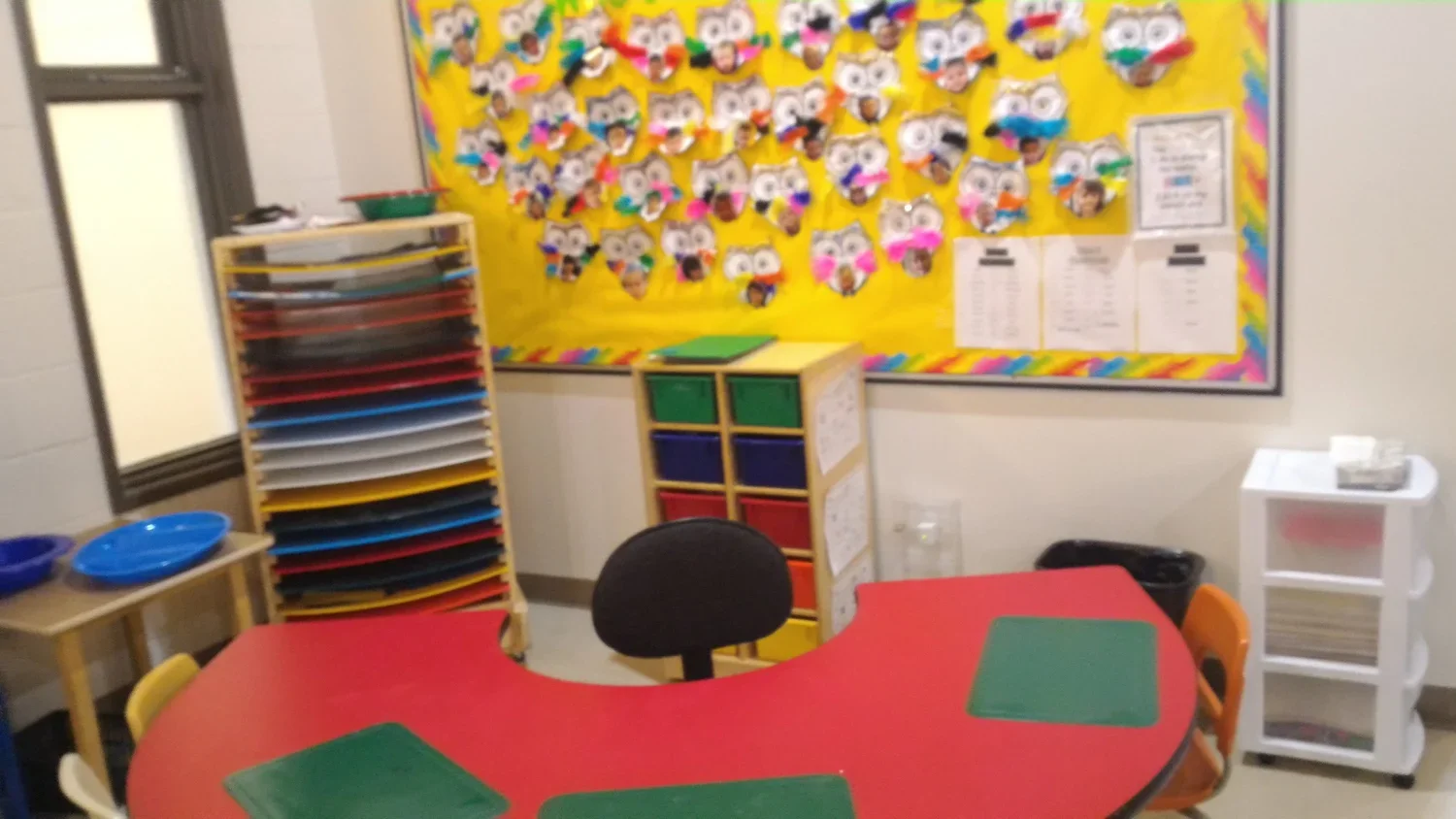 Classroom corner with a red semi-circular table, black chair, storage cubbies with colored bins, a wall decorated with owl crafts, and various classroom supplies.