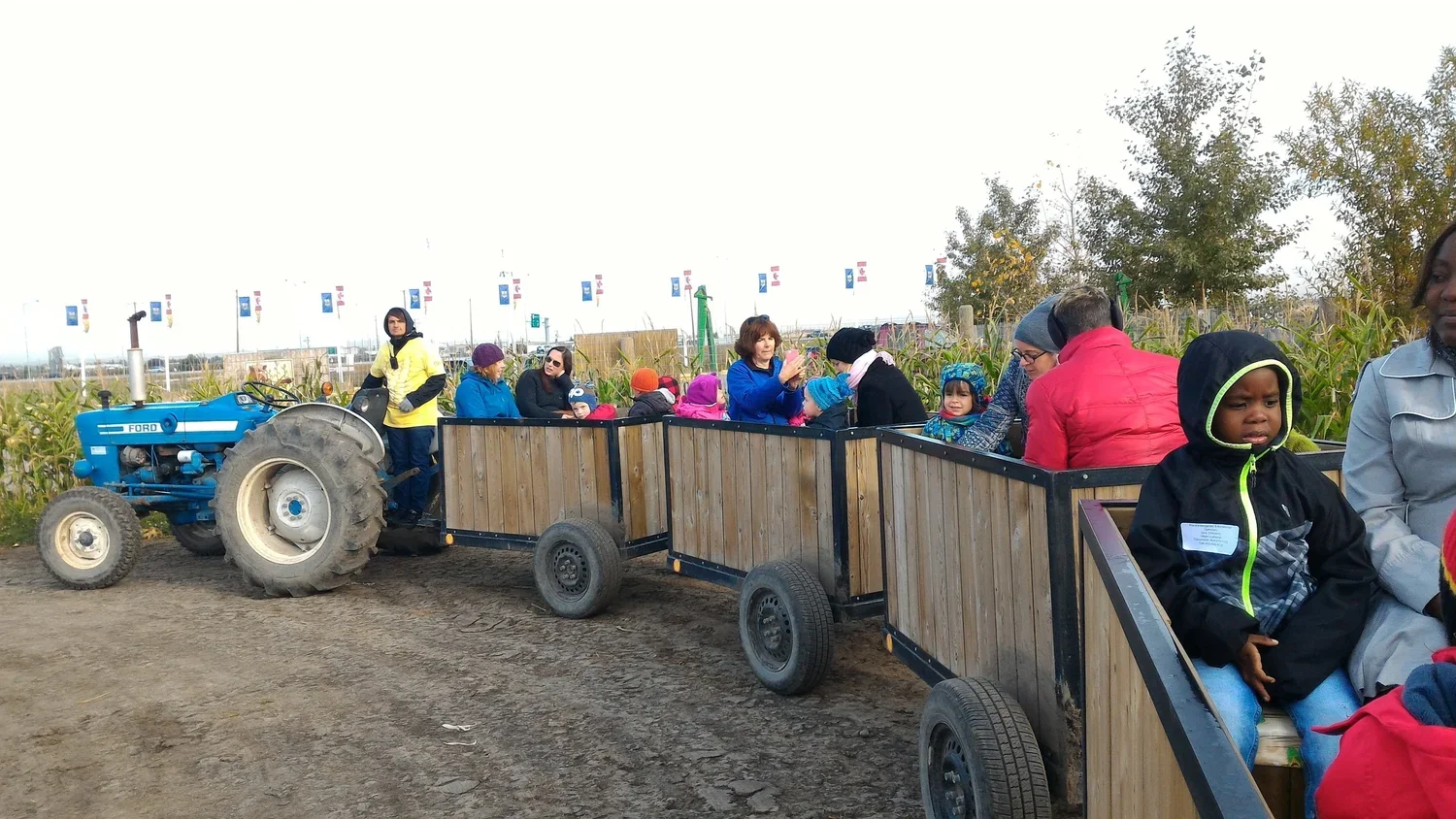 Children and adults riding in a hayride wagon pulled by a blue tractor at a farm, with cornfields in the background.