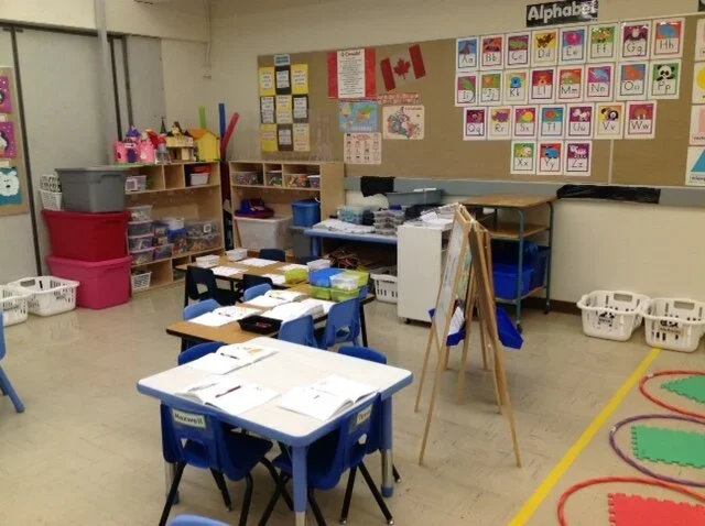 A classroom with desks, shelves filled with supplies, colorful educational posters on the wall, and small chairs for students.