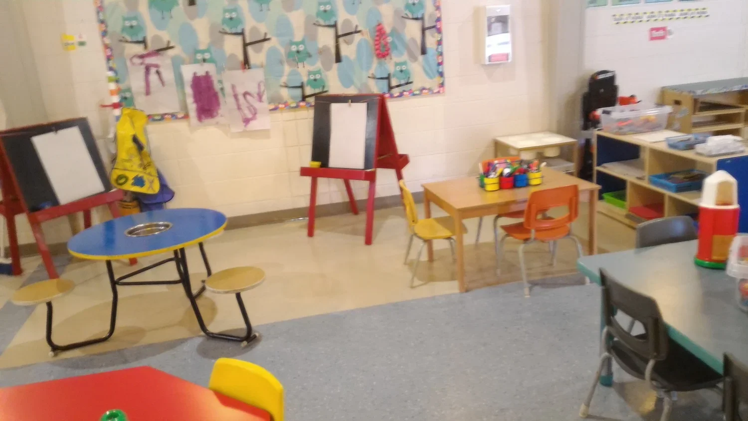 A preschool classroom with small tables and chairs, a colorful wall display, and various educational and art supplies.