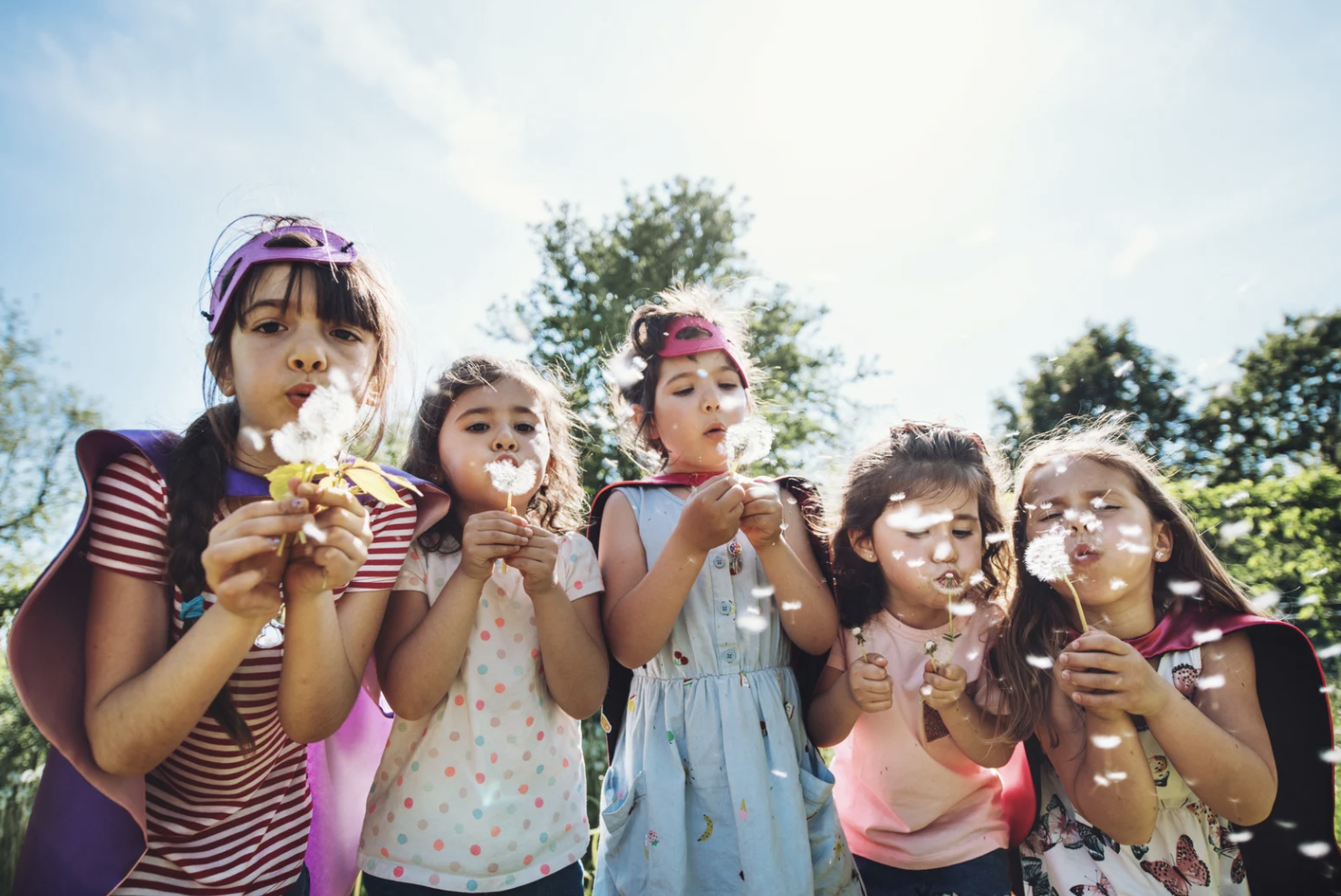 Five young girls outdoors blowing dandelion seeds on a sunny day with trees in the background.