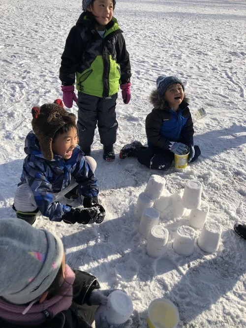 Three children in winter clothing playing in the snow, building a snow castle with cups and buckets, while smiling and enjoying the outdoor winter activity.