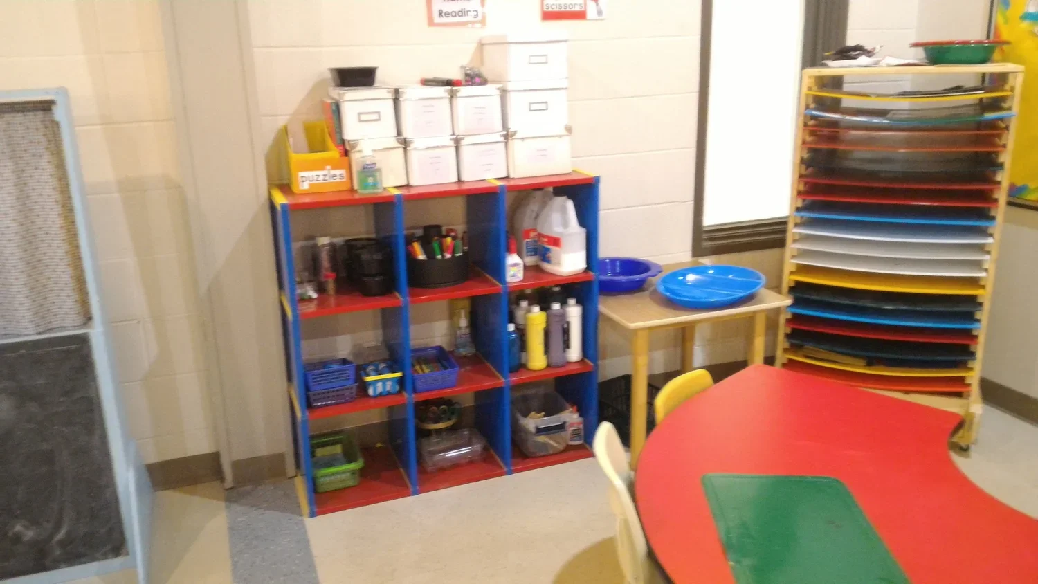 Colorful classroom corner with storage shelves, a small table with a blue tray, a red and yellow table with chairs, and a stack of plastic trays of various colors.