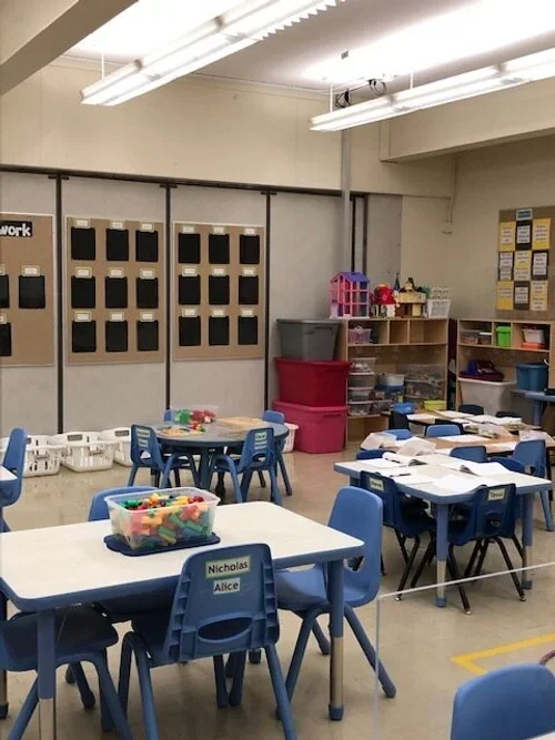 Classroom with small tables and blue chairs, storage bins, and educational materials.