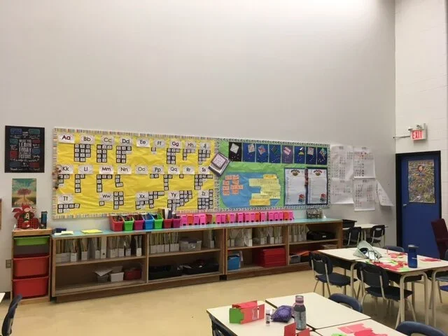 Classroom with alphabet and number decorations, colorful storage bins, a bookshelf, tables with supplies, and a door with a poster.