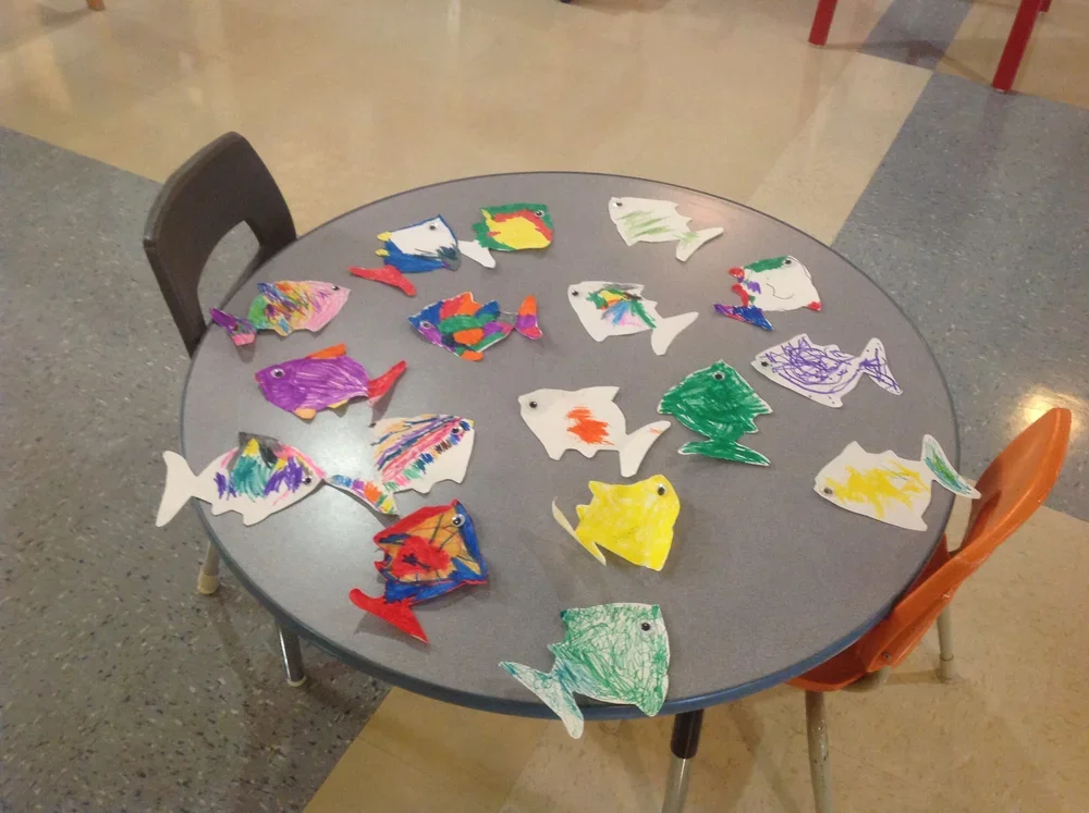 Colorful paper fish cutouts decorated with crayons on a round gray table in a classroom setting.
