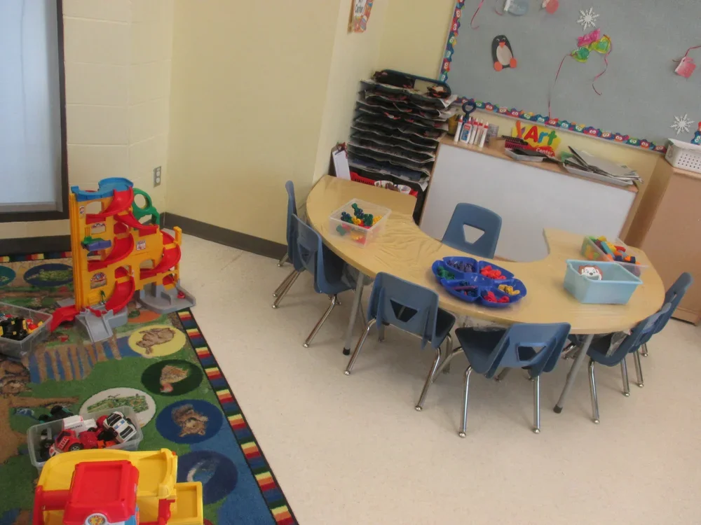 A small classroom with a table surrounded by six blue chairs, toys and building blocks on the table, a colorful rug with animal pictures on the floor, storage bins and shelves with papers and toys, and a bulletin board decorated with penguin and snow