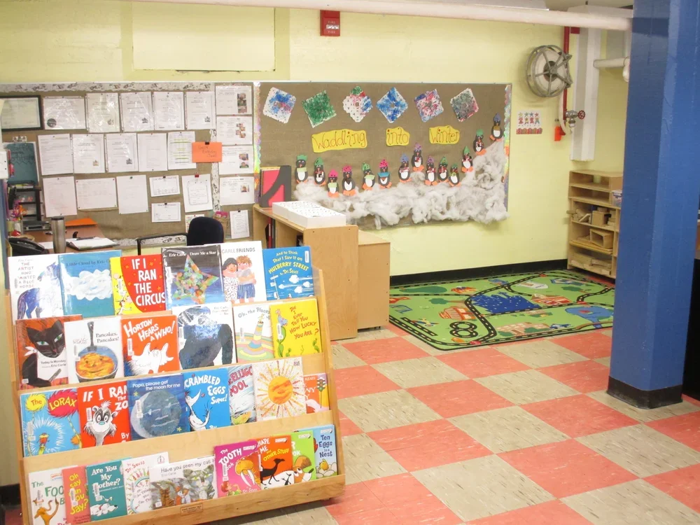 A classroom library corner with a bookshelf filled with children's books, a wall display with penguin decorations, and a colorful rug with a train track pattern.