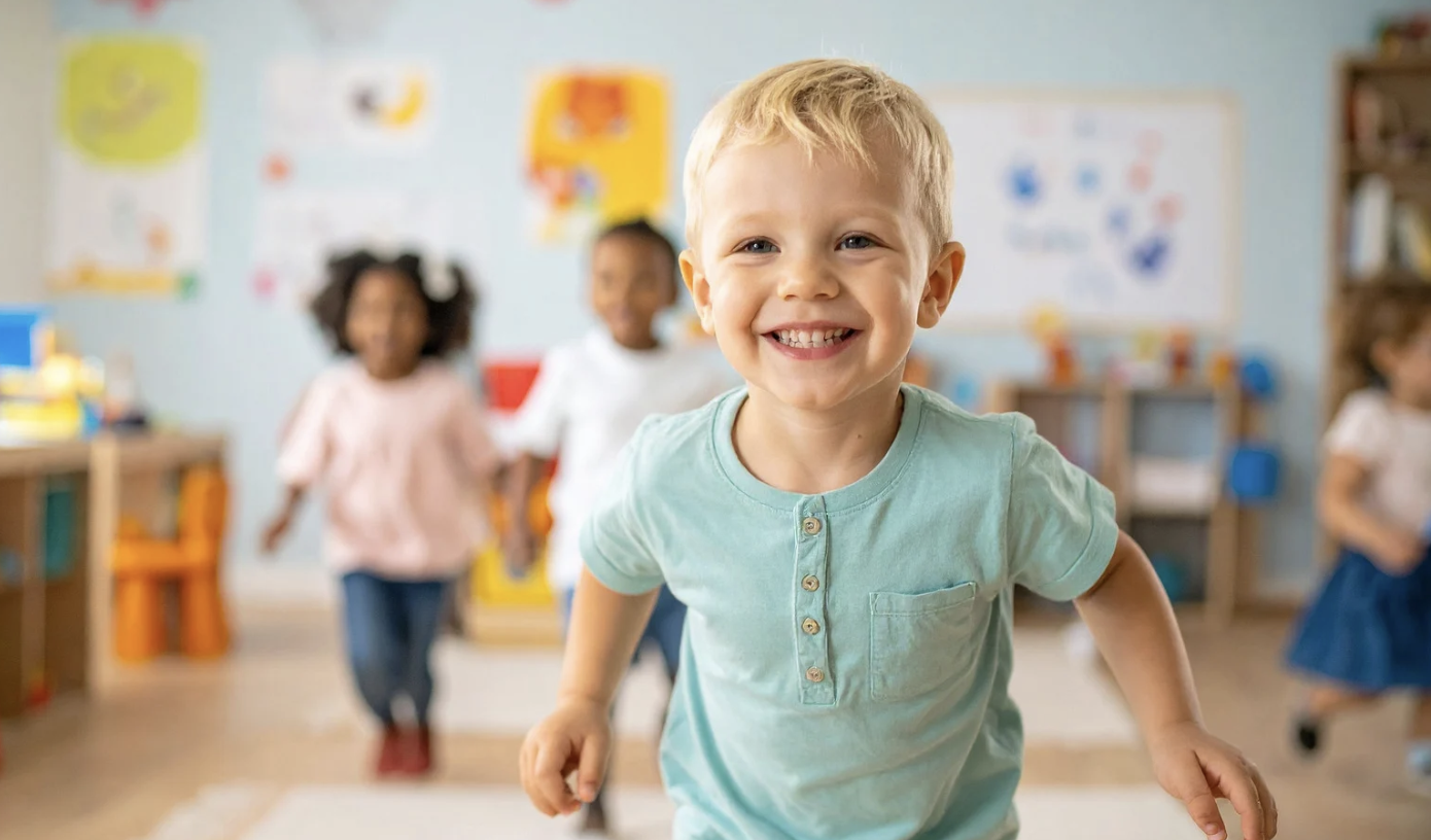 Young boy smiling and running in a classroom with other children in the background.