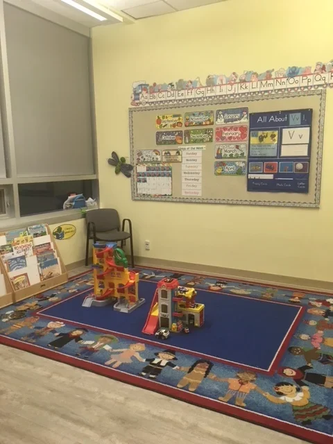Classroom with a blue area rug featuring cartoon children, a small black chair, educational posters on the yellow wall, and toy building blocks arranged for play.