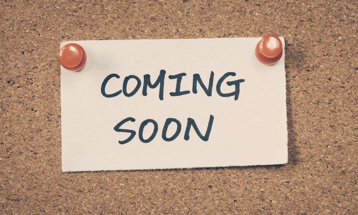 A white note pinned to a corkboard with orange push pins that reads 'Coming Soon' in black handwritten font.