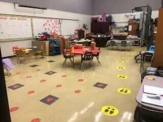 Empty classroom with chairs arranged around tables, educational posters on the walls, and yellow social distancing markers on the floor.