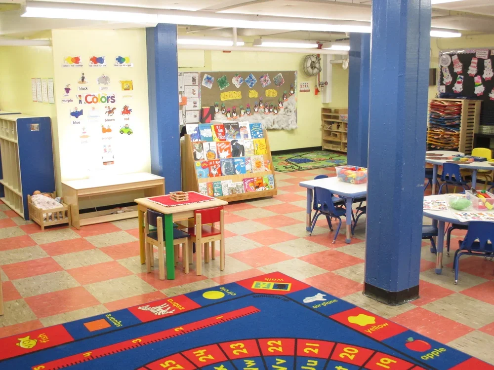 A colorful preschool classroom with educational posters, books, tables, and chairs, alongside a blue and yellow rug with numbers and shapes.