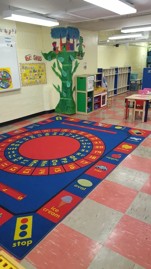 Classroom with a colorful educational rug featuring numbers, shapes, and words, and a mural of a green tree with a castle on top on the wall.