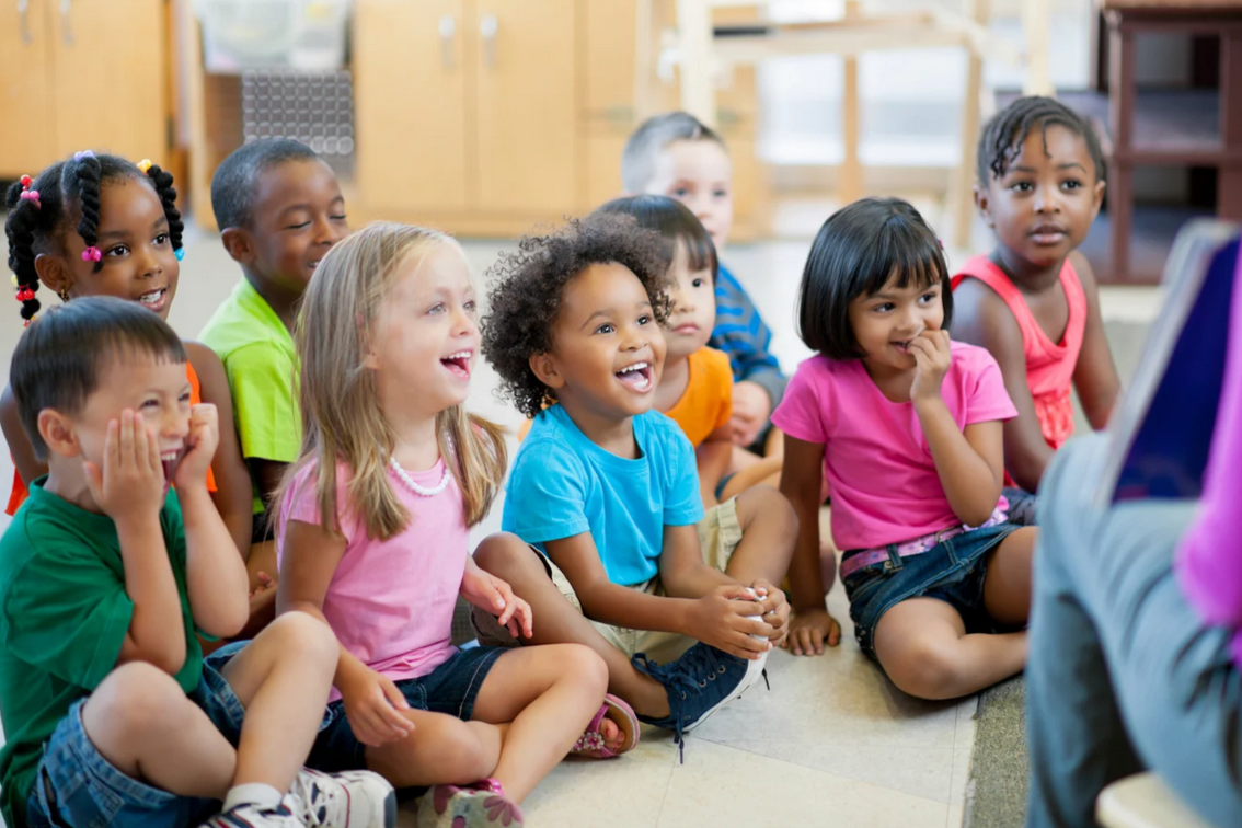 A group of young children sitting on the floor, smiling and laughing, watching someone in front of them in a classroom or childcare setting.