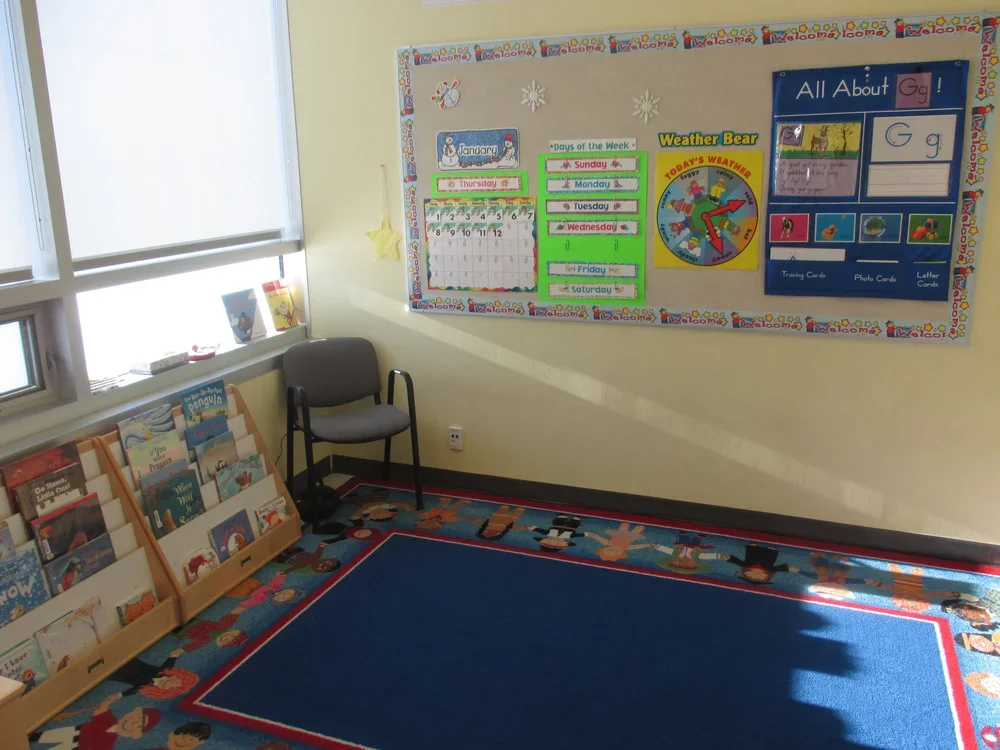 A classroom corner with a reading nook, a black chair, a bookshelf filled with children's books, and a wall decorated with educational posters about days of the week, weather, and the alphabet.