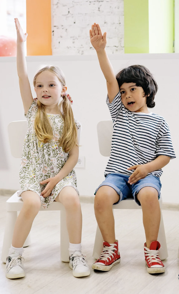 Two children, a girl and a boy, sitting on chairs in a room with colorful walls, raising their hands as if answering a question.