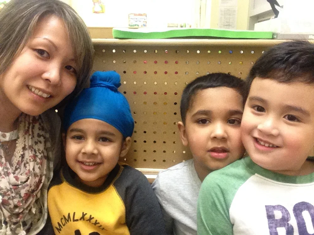 A woman with light brown hair taking a selfie with four children of diverse ethnic backgrounds in a classroom or similar setting.