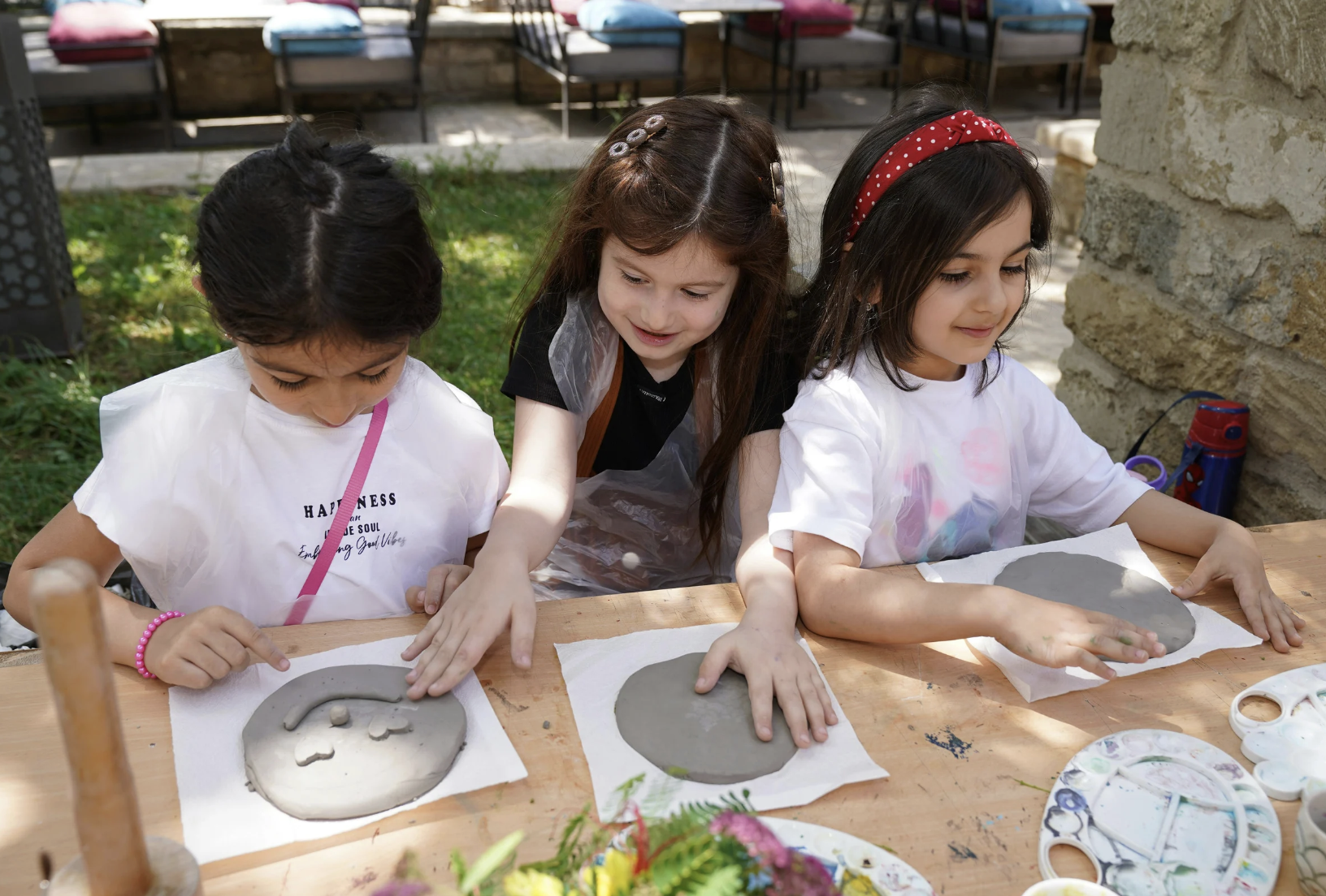 Three young girls are engaging in outdoor clay art activity at a wooden table, using their hands to mold gray clay into shapes.