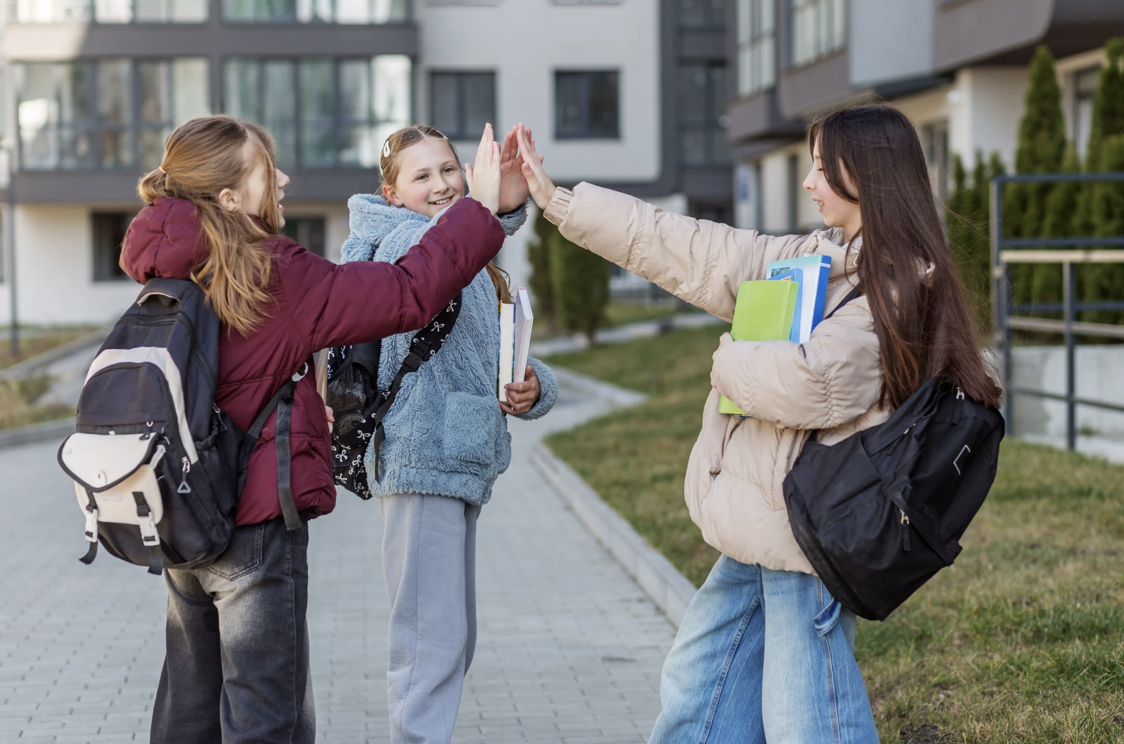Three children giving each other a high five outdoors on a school sidewalk, carrying backpacks and books, with modern apartment buildings in the background.