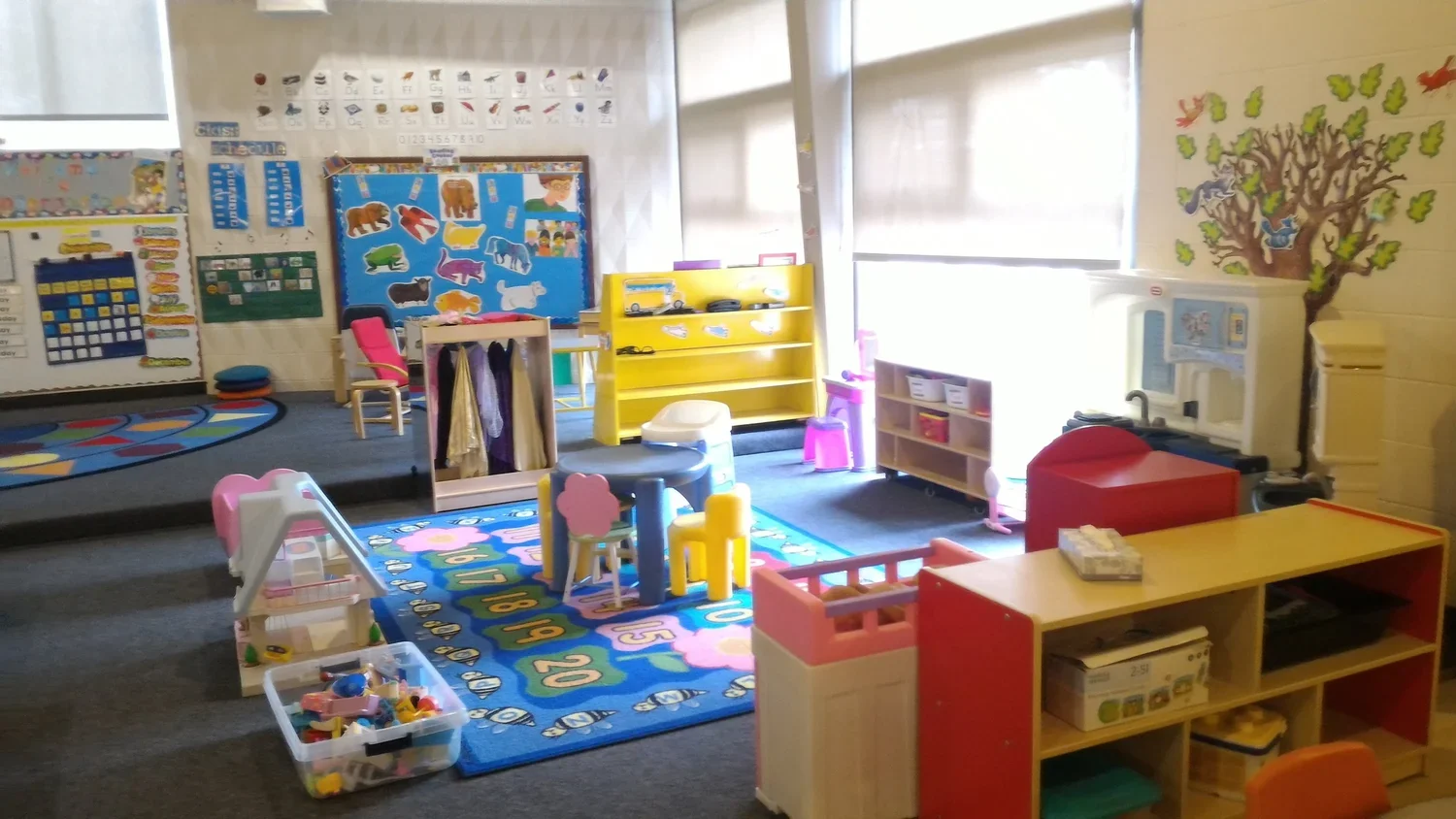 A preschool classroom with colorful furniture, educational posters, a play kitchen, and various toys and rugs for children to play and learn.