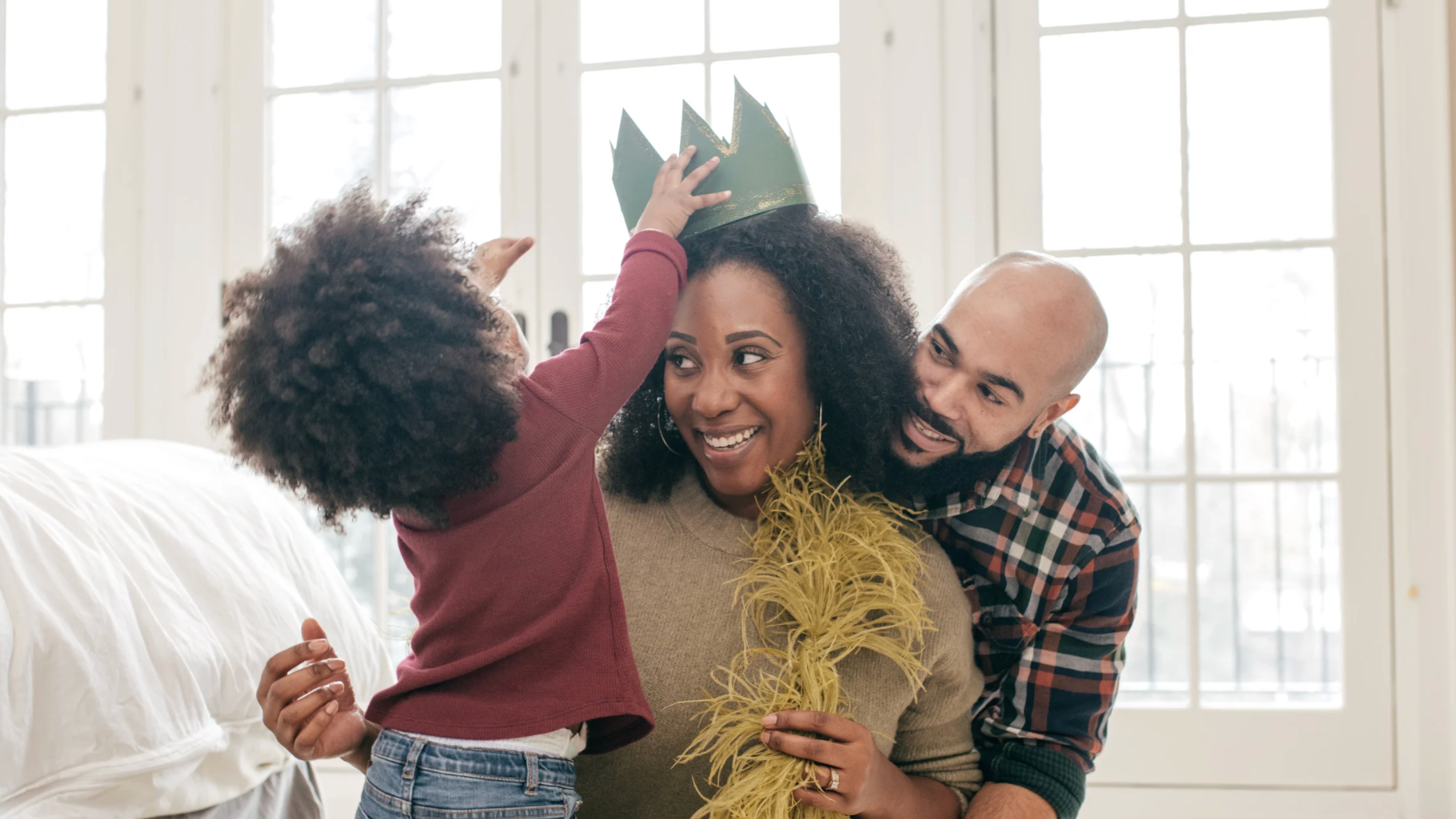 A family celebrating a birthday indoors with a woman receiving a green paper crown from a child, while a man smiles beside them. The woman is holding yellow feathered decorations, and there are large windows in the background.