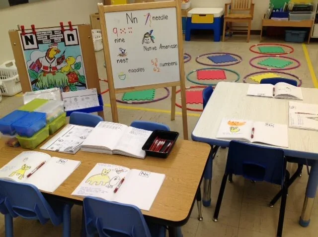 Classroom with tables, open books, and a whiteboard displaying the letter 'Nn' and related words. Coloring pages and educational materials are visible, along with colorful circular rugs on the floor.
