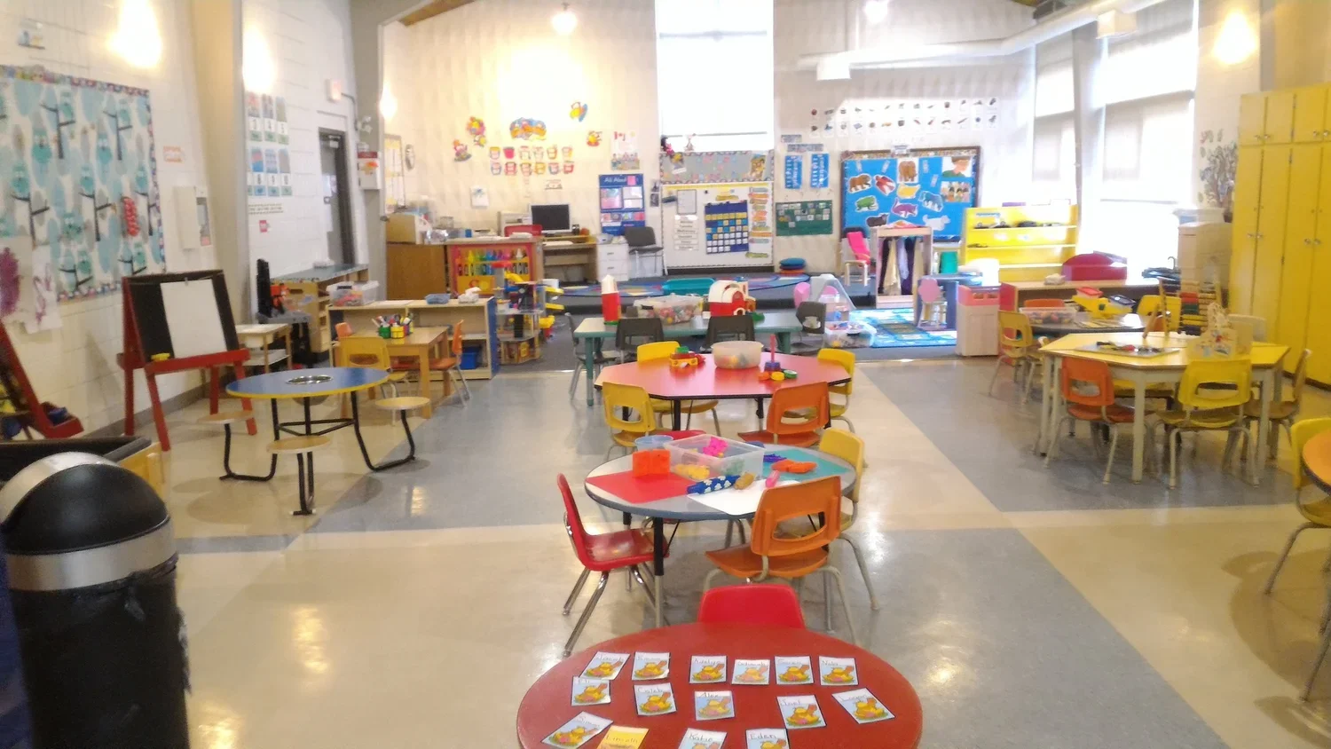 A colorful classroom with round tables, small chairs, educational posters, toys, and storage lockers, likely for young children or preschoolers.