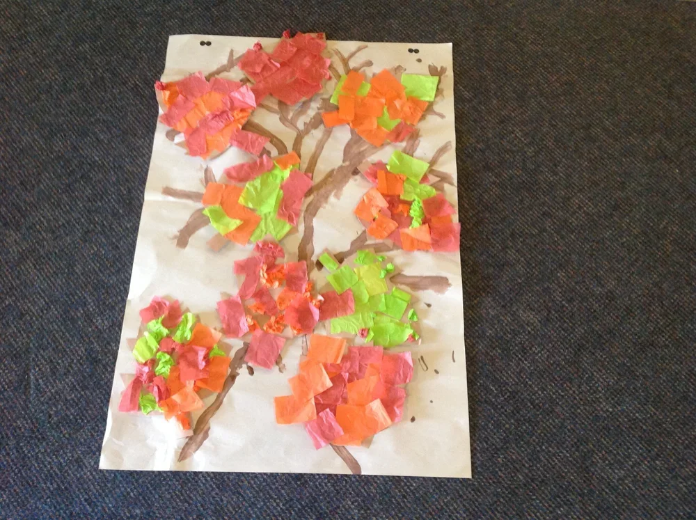 A handcrafted collage of a tree on white paper, with torn colorful tissue paper in pink, orange, and green forming the leaves, laid on a dark fabric surface.