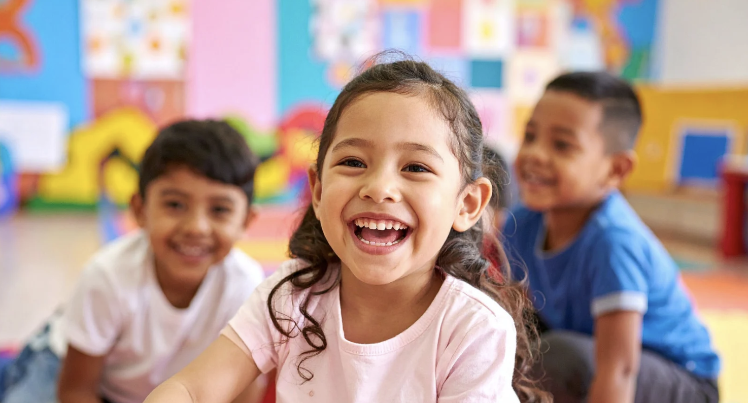 Smiling young girl with curly hair and two boys in a colorful classroom setting.
