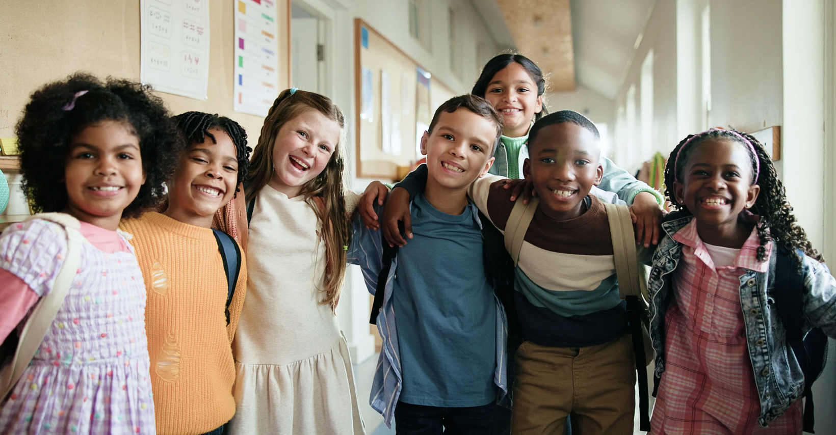 Group of eight diverse elementary school children smiling and posing together in a hallway.