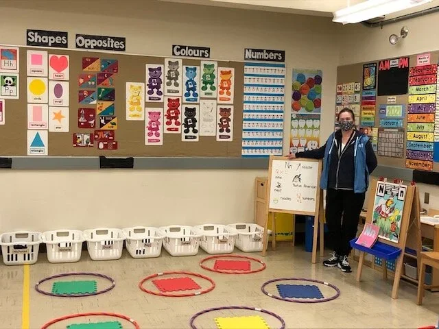 A classroom with educational posters on the wall about shapes, opposites, colors, and numbers. A teacher or student wearing a face mask stands next to a whiteboard and a reading bookshelf. Several baskets are on the floor, each inside a colorful hula