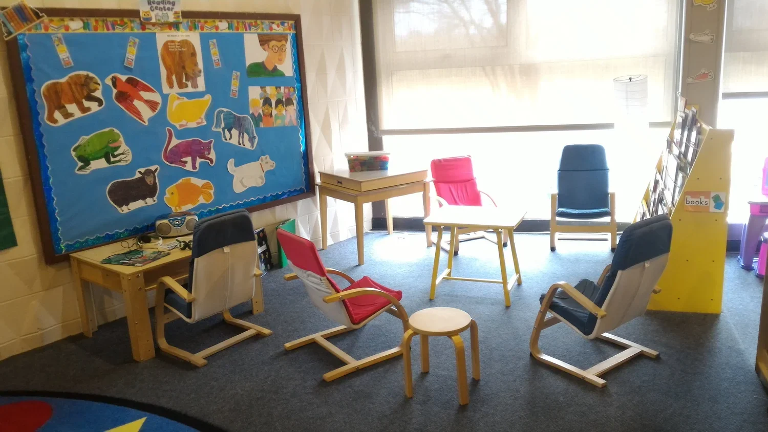 A quiet reading corner in a classroom with chairs, a small table, a bulletin board with animal pictures, and large window providing natural light.