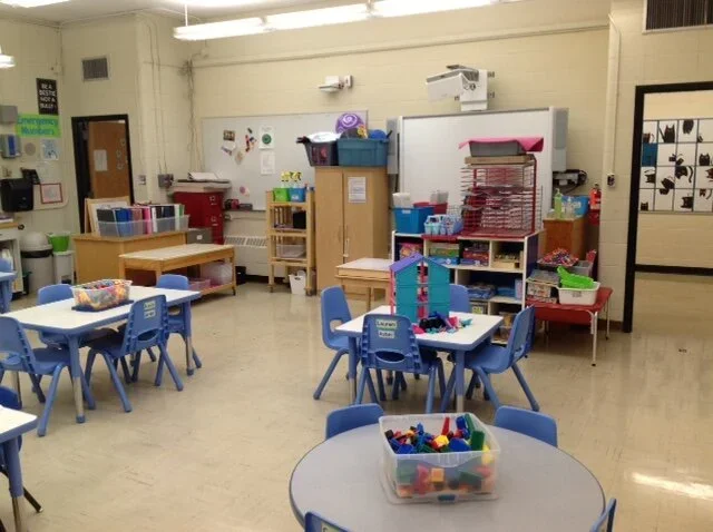 Classroom with small blue tables and chairs, educational supplies and storage shelves, and a whiteboard.