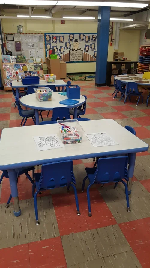 Empty classroom with blue tables and chairs, coloring pages, markers, books, and a colorful bulletin board with student artwork.