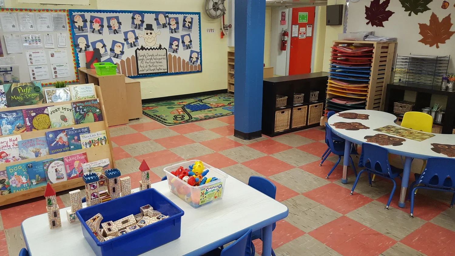 A colorful classroom with books, educational toys, and furniture for young children. There are tables with chairs, a book display, and a bulletin board with children's artwork.