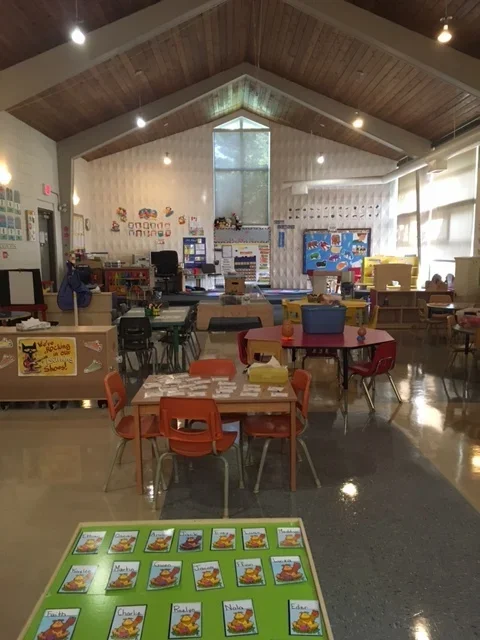 A spacious classroom with a high, wooden, pitched ceiling and large windows on the right side allowing natural light. In the foreground, there's a table with various cards featuring a cartoon animal, likely a classroom activity. Multiple small tables