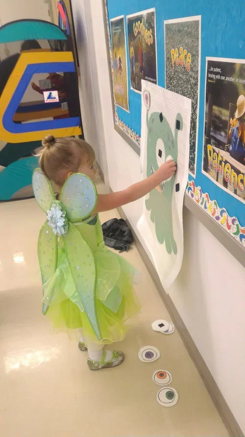 A young girl dressed as a fairy with green butterfly wings and a green dress is interacting with a large bear-shaped paper on a wall. She appears to be adding or pointing at something on the paper. There are posters and artwork on the blue wall next 