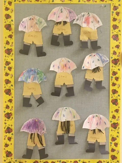 Childcraft craft display featuring paper umbrellas above paper cutouts of legs wearing shoes. Each craft has a child's name written on it, including Emile, Tiffany, Albertina, Yuragi, Lewis, Mei, and others. The display is framed with a yellow border