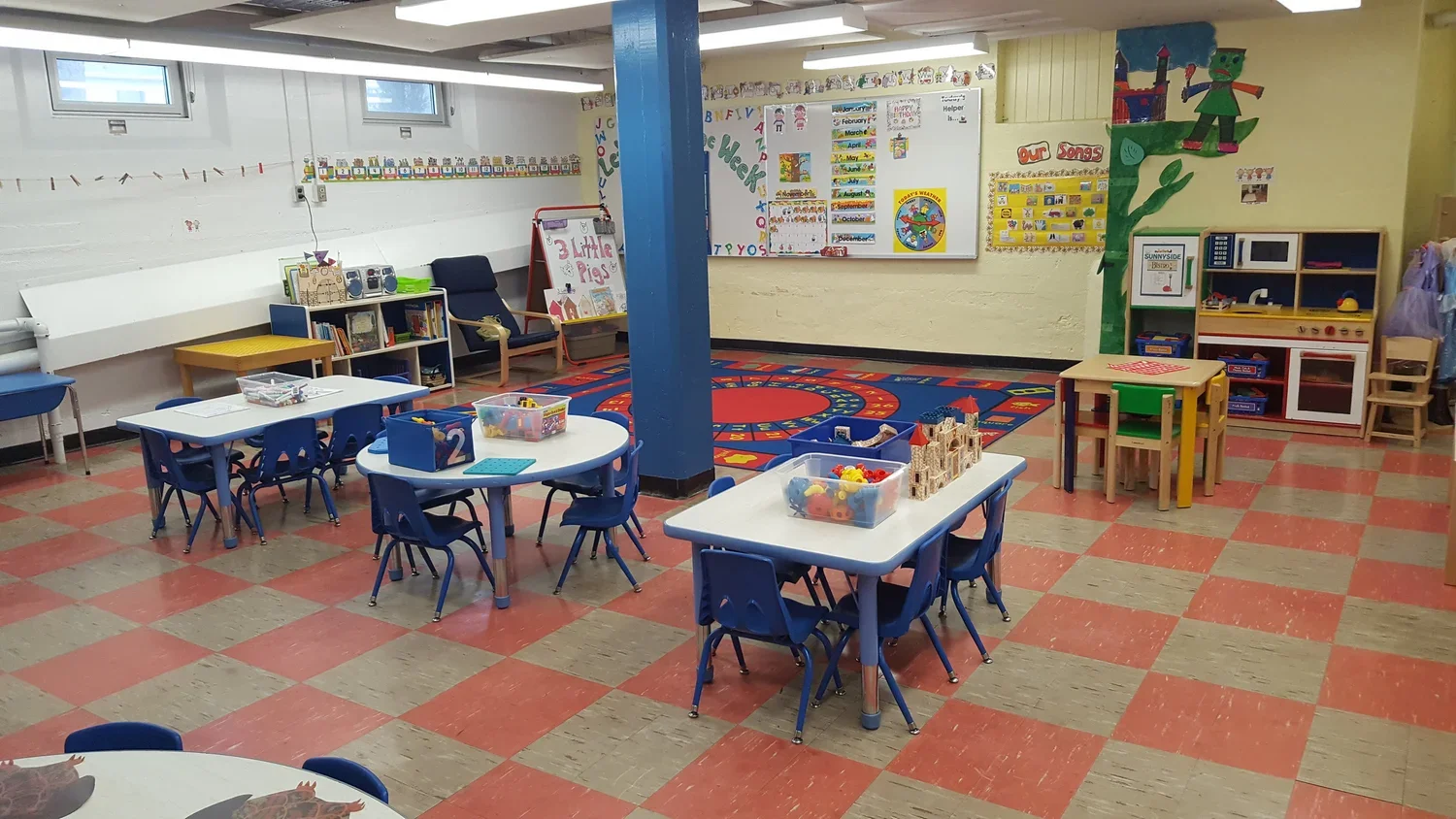A colorful classroom with small blue tables and chairs, educational toys, posters, and a play kitchen area.