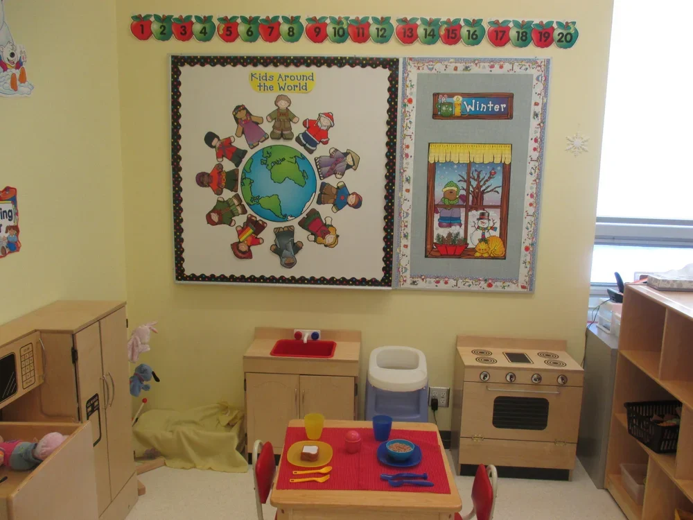 Classroom corner with a bulletin board featuring a poster titled 'Kids Around the World' and a winter-themed display. There is a table set with toys and utensils, small kitchen furniture, and a row of numbered apple decorations above the bulletin boa