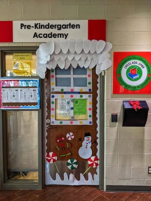 Decorated door with a Christmas theme in a preschool, labeled 'Pre-Kindergarten Academy'. The door has foam ice cream cone decorations, paper snowman, gingerbread man, and candy cane cutouts, with white cloud-like trim at the top.