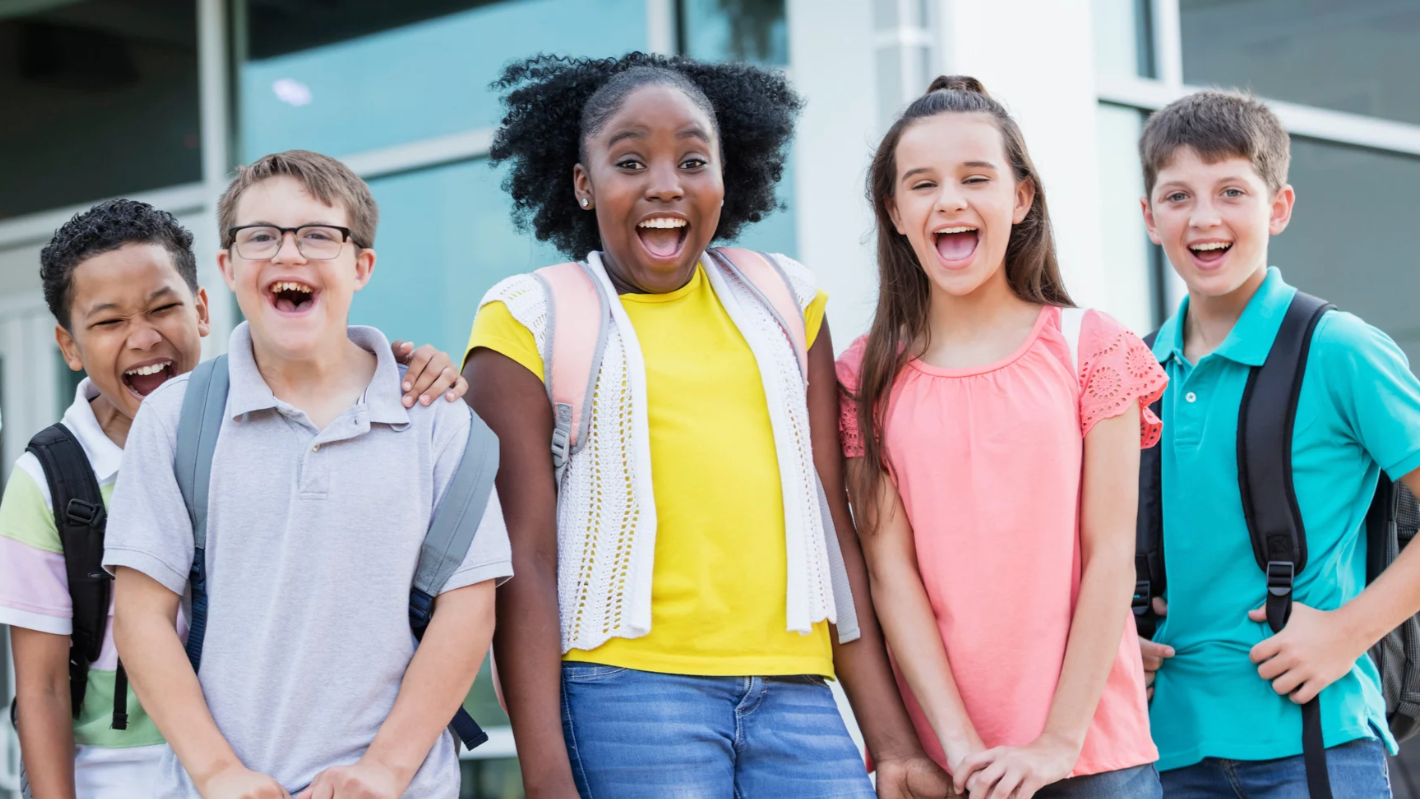 Group of five happy diverse schoolchildren standing outside, smiling and laughing.