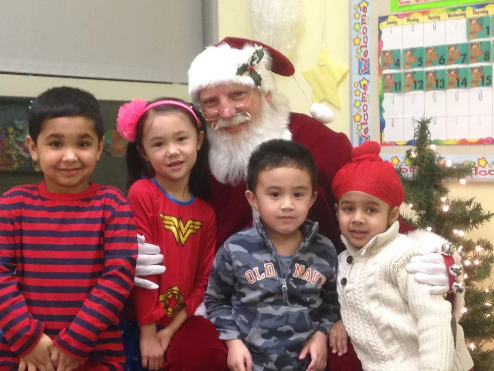 Santa Claus with four children posing in a classroom decorated for Christmas, including a small Christmas tree and colorful calendar on the wall.