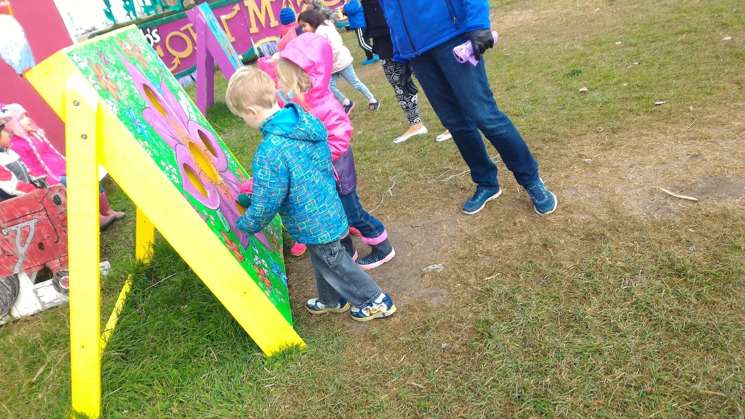 Children playing a ring toss game outdoors on grass, supervised by an adult, with colorful painted targets and nearby seats.
