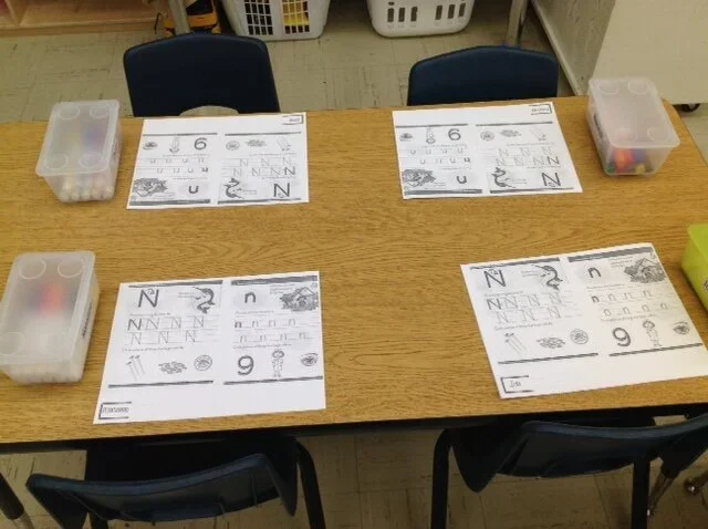 Four tables, each with a worksheet, a plastic container, and some chairs around them in a classroom setting.