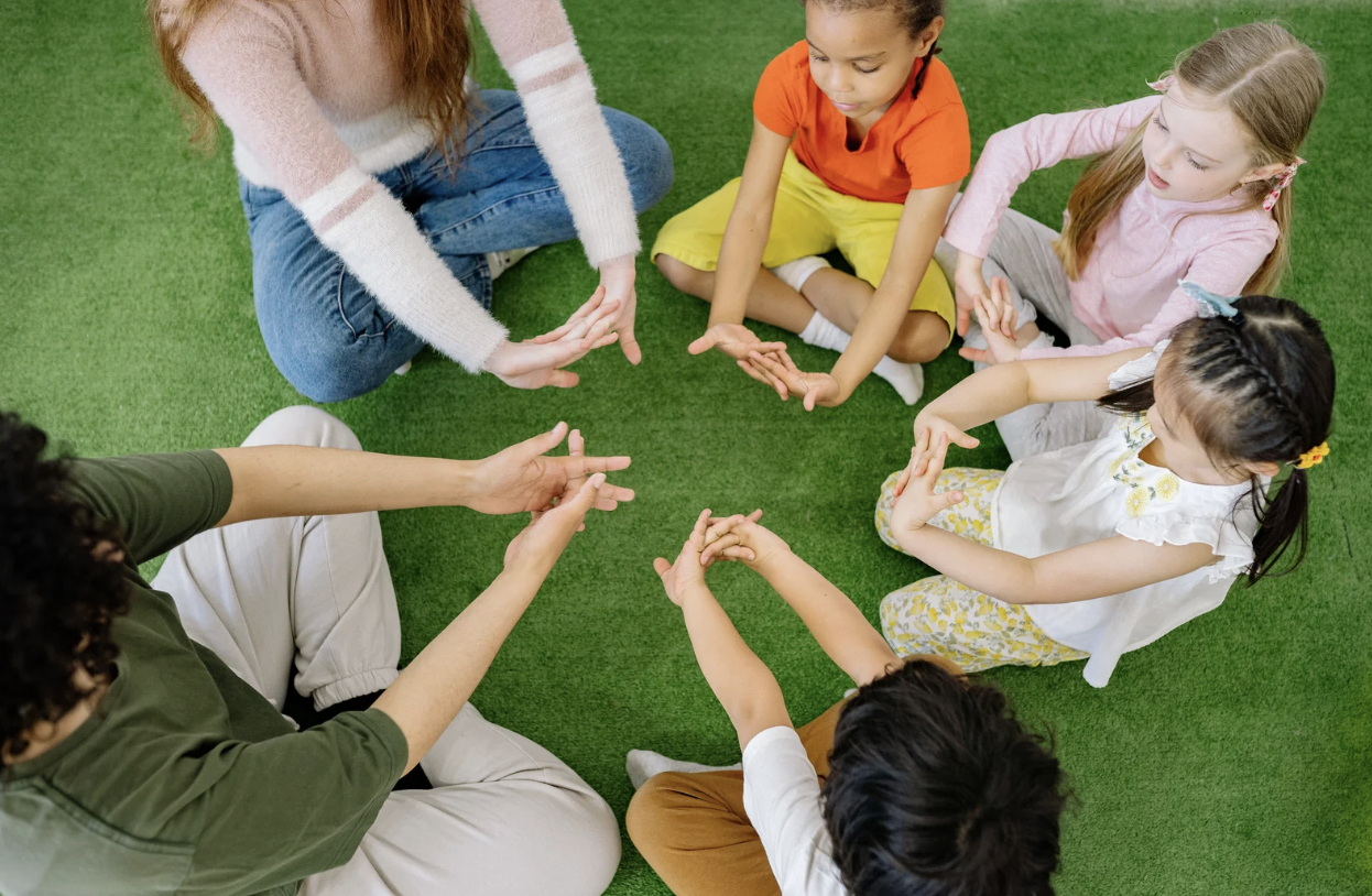 Group of children and an adult sitting on green carpet in a circle, engaging in a group activity.